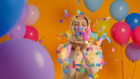Studio-Portrait-Of-Woman-In-Hijab-Wearing-Birthday-Queen-Headband-Celebrating-Blowing-Paper-Party-Confetti-To-Camera